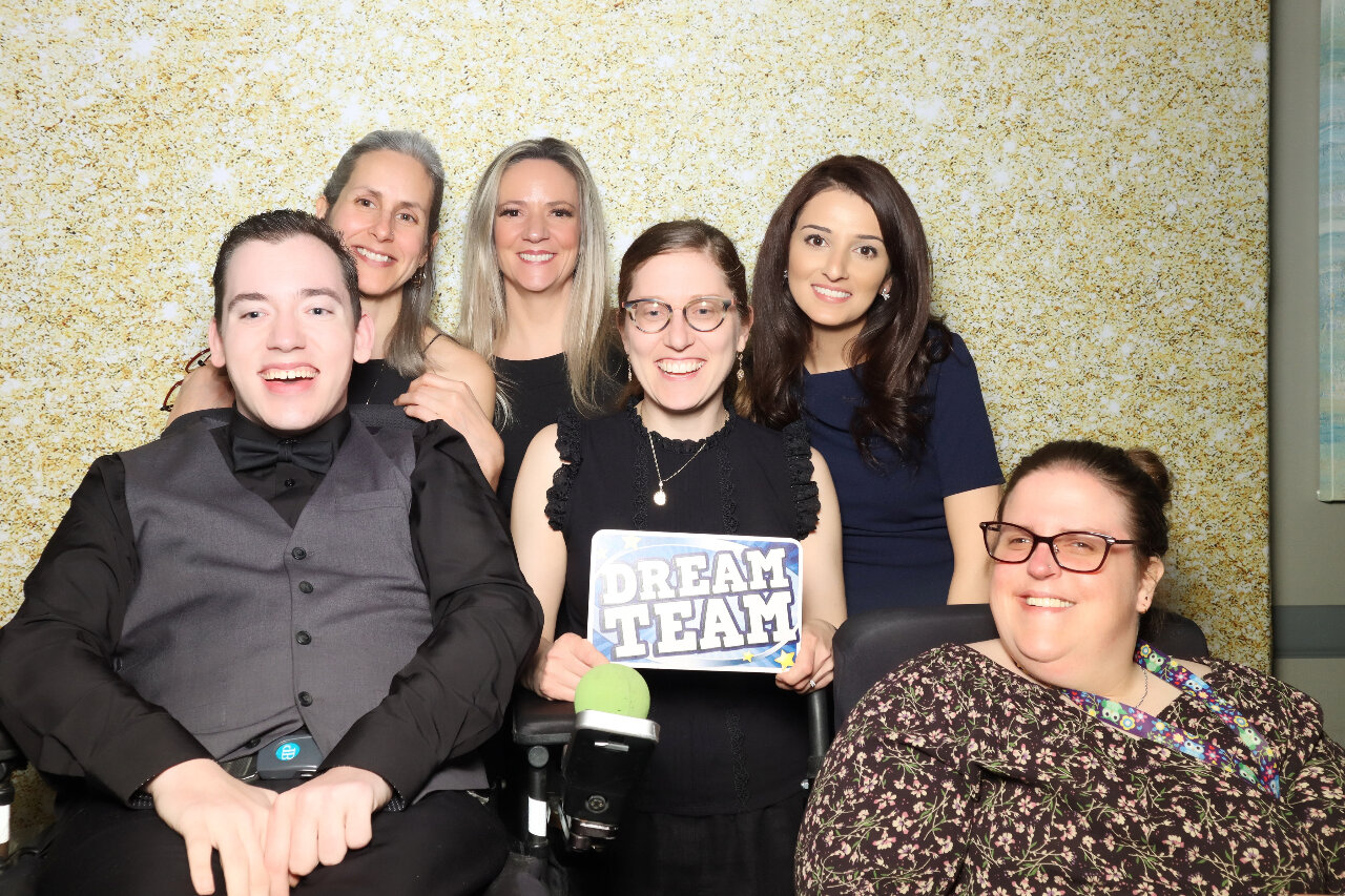 Seven WRFN staff posing together, From left to right, Keenan O'Donoghue, Leah Bowman, Lisa Cook, Cristina Sanger, Oula Almadhoun  and Carmen Sutherland are in front of a glittery gold backdrop. Keenan and Carmen are using wheelchairs, Cristina in the center holds a sign that reads "Dream Team." Everyone is smiling warmly at the camera.